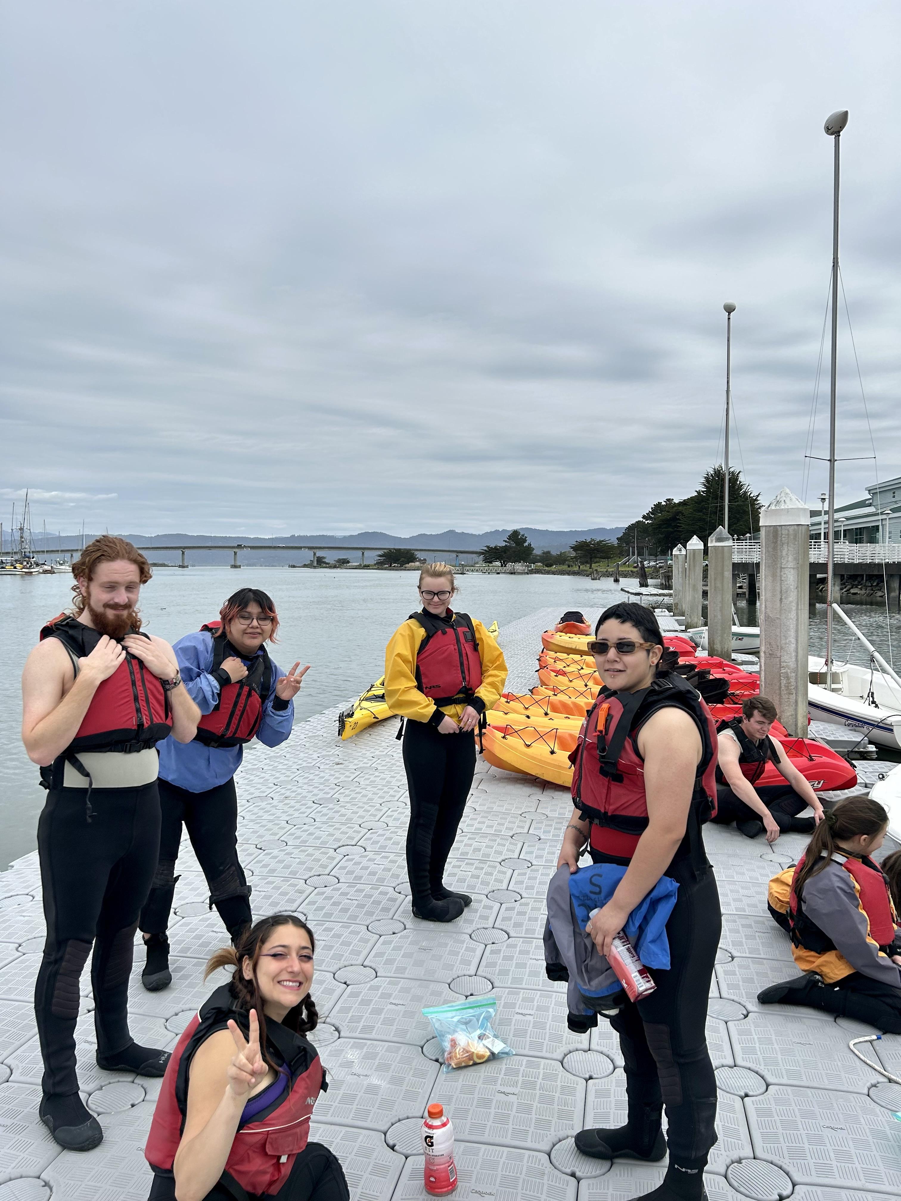 LEAP volunteers and students from Orick School stand on the dock at the Humboldt Bay Aquatic Center wearing wet suits and life vests. They ware about to go kayaking