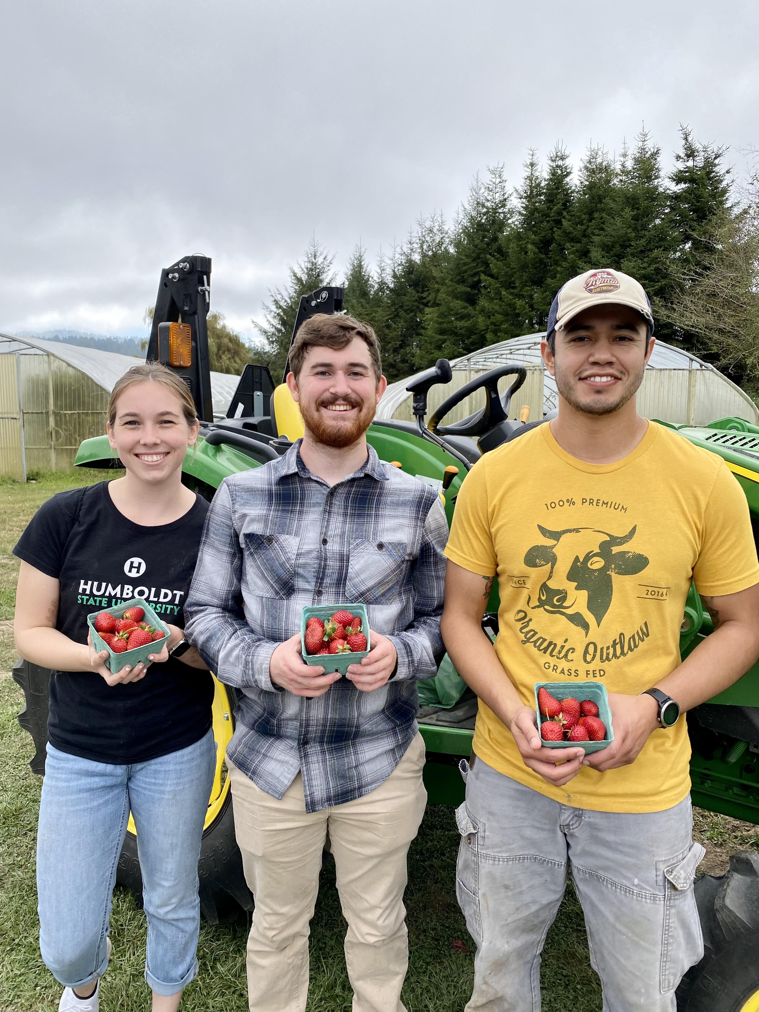 Three Cal Poly Humboldt students stand in front of a tractor holding baskets of strawberries